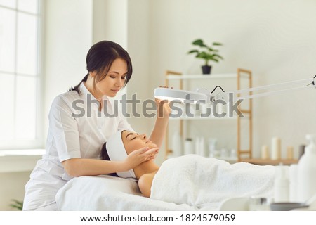Young smiling dermatologist looking at young womans face during skin examination under lamp in beauty salon. Professional skin checking concept Royalty-Free Stock Photo #1824579059