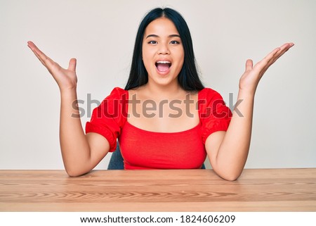 Young beautiful asian girl wearing casual clothes sitting on the table celebrating victory with happy smile and winner expression with raised hands 
