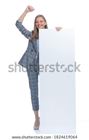 enthisiastic young woman in blue checkered suit holding arm in the air and cheering, presenting empty board and smiling, sitting isolated on white background, full body