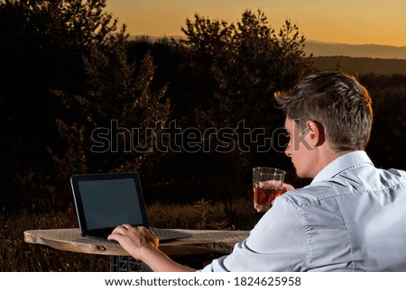 A man in a white shirt works at a laptop. Work on nature background. Sunset in summer. The concept of remote work and freelance. Mini office on an old wooden board. He drinks whiskey from a glass.