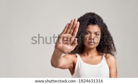 Portrait of young african american woman with curly hair wearing white shirt having serious confident look while making stop gesture isolated over grey background. Front view. Focus on hand