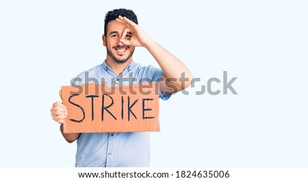 Young hispanic man holding strike banner cardboard smiling happy doing ok sign with hand on eye looking through fingers 