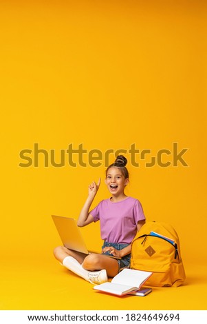 happy schoolgirl with backpack, laptop and books is sitting on yellow background in the Studio. The child shows, a sign of approval, a finger up. Back to school. copy space. Royalty-Free Stock Photo #1824649694