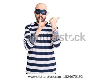 Young handsome man wearing burglar mask asking to be quiet with finger on lips pointing with hand to the side. silence and secret concept. 