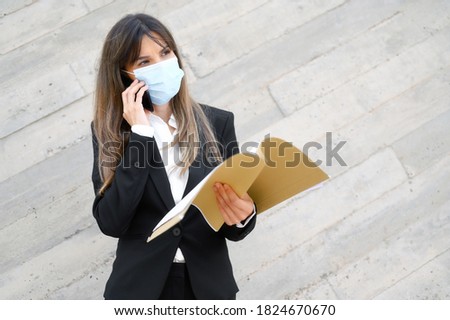 Portrait of a cheerful confident businesswoman in business suit wears protective face mask to prevent COVID-19, holds document file and mobile phone at office building. High quality photo
