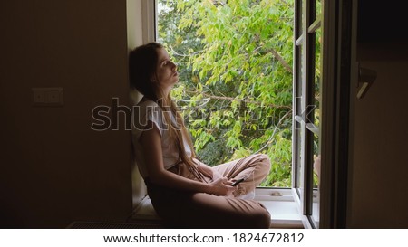 A young, Caucasian, pretty blonde with long hair sits on the windowsill of an open window in a lotus position and listens to music on wired headphones from her smartphone. Royalty-Free Stock Photo #1824672812