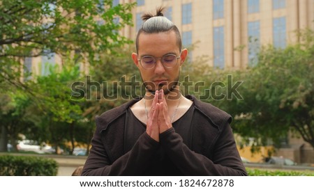 Young caucasian charismatic brunette man with a ponytail on his hair and round glasses makes a namaste gesture, crossing his arms on his chest. Positive emotions. Non-verbal gestures. Royalty-Free Stock Photo #1824672878