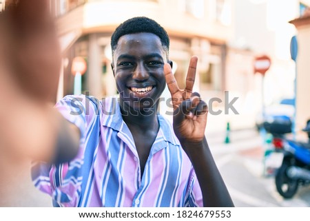 Young african american man making selfie by the camera doing victory sign with fingers at street of city.