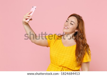 Smiling beautiful young redhead plus size body positive chubby overweight woman in yellow dress posing doing selfie shot on mobile phone isolated on pastel pink color wall background studio portrait
