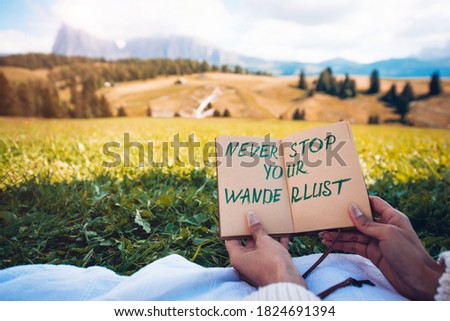 Hands holding page text "Never stop your wanderlust" with nature hill landscape on the background. Travels and motivational concept