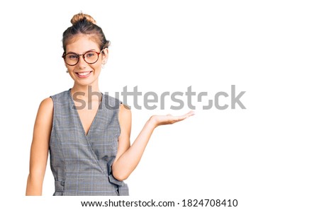 Beautiful caucasian woman with blonde hair wearing business clothes and glasses smiling cheerful presenting and pointing with palm of hand looking at the camera. 