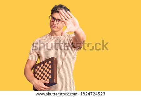 Young handsome man holding chess with open hand doing stop sign with serious and confident expression, defense gesture 