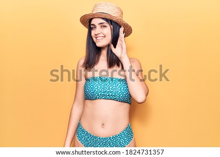 Young beautiful girl wearing bikini and summer hat smiling with hand over ear listening an hearing to rumor or gossip. deafness concept. 