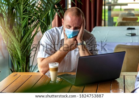 Shot of concerned young man using laptop at cafe wearing face mask,protecting himself from coronavirus.Caucasian businessman protected with a face mask working on his laptop at the local coffee shop.