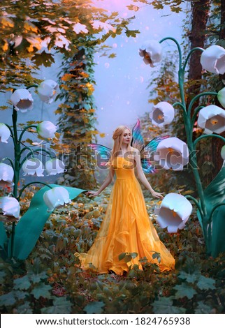 A fantasy woman forest fairy. Fashion model in yellow dress with butterfly wings walks in autumn nature. Large flowers scenery decor white lily of the valley. Orange leaves of tree blue mystical fog