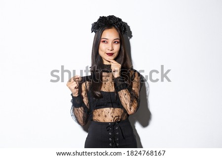 Image of smiling beautiful asian woman in gothic lace dress and wreath, thinking while holding credit card, standing over white background