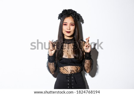 Image of hopeful asian woman in gothic lace dress and wreath making wish, cross fingers for good luck, looking wishful at camera, standing over white background