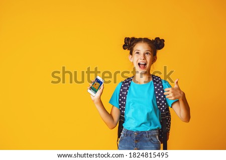 Cute positive schoolgirl 10 years old in casual clothes with backpack, sitting on yellow background, in Studio, holding mobile phone. Dependence on gadgets and Internet. E-education. Copy space. Royalty-Free Stock Photo #1824815495