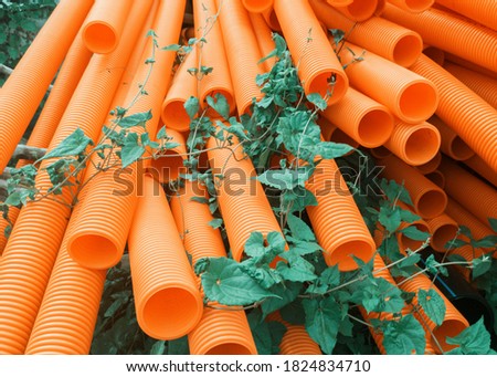 Closeup of stacked orange corrugated PVC plastic pipes, lying on ground in construction site. Few green weeds are seen growing around them.