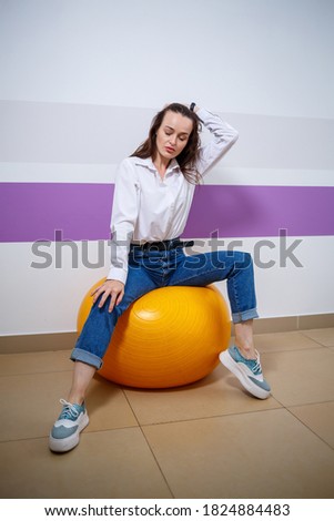 A girl of European appearance with different emotions on her face sits on a ball for a gymnast. Attractive young brunette woman smiling and fooling around
