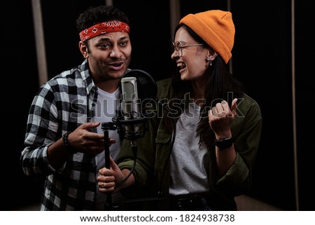 Portrait of young man and woman, duet singing into a condenser microphone while recording a song in a professional studio