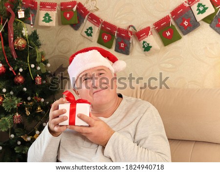 Happy senior man in a Santa hat is holding a gift in front of a Christmas tree. The concept of New year and a happy Christmas