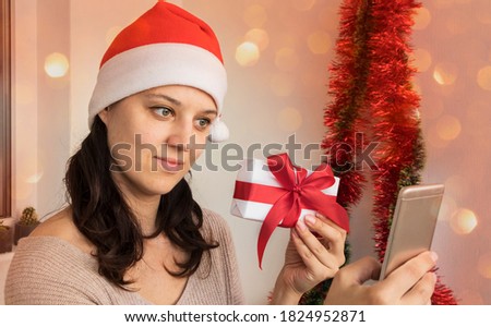 Woman in a Santa hat holds a gift box and shows it to a smartphone. Tinted photo with bokeh added. Merry christmas and happy new year greetings online