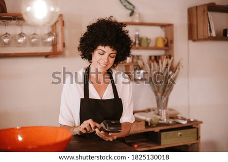 Smiling female cafe owner standing working with a card terminal. Woman with apron standing behind the bar counter of a modern restaurant. Concept small business owner, young entrepreneur.