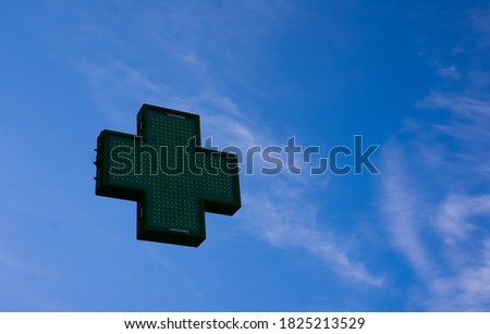 From a low angle you can see a green pharmacy cross floating on a blue sky with some light clouds