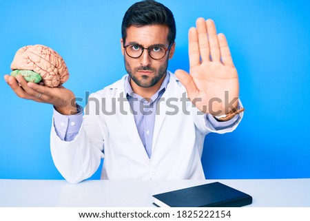 Young hispanic man sitting at the table wearing doctor coat holding brain with open hand doing stop sign with serious and confident expression, defense gesture 