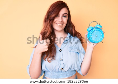 Young beautiful woman holding alarm clock smiling happy and positive, thumb up doing excellent and approval sign 