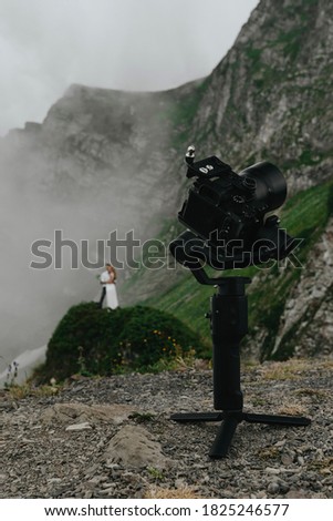 stabilizer with a camera standing in the mountains in cloudy weather, behind him is the groom and the bride in a wedding dress, wedding photography, moody colors