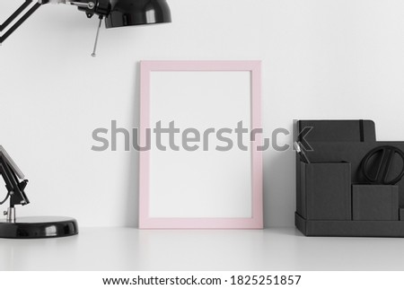 Pink frame mockup with a lamp and workspace accessories on a white table. Portrait orientation.