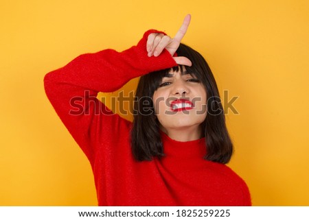 Caucasian brunette woman wearing red casual sweater isolated over yellow background making fun of people with fingers on forehead doing loser gesture mocking and insulting.