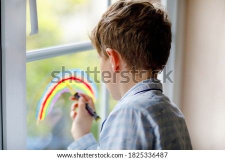 Lovely school kid boy in pajamas painting rainbow with colorful window color during pandemic coronavirus quarantine. Children painting rainbows around the world with the words Let's all be well.