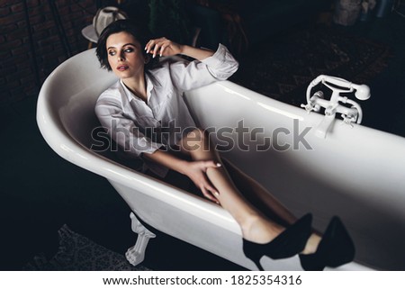 Portrait of a beautiful brunette lying in an empty bathroom holding legs up, wearing a white shirt and black shoes on red brick wall background.