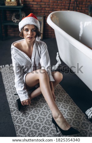 A young brunette posing on the floor near the bath in Сhristmas hat, white shirt and black high heels.