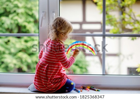 Adoralbe little toddler girl with rainbow painted with colorful window color during pandemic coronavirus quarantine. Child painting rainbows and hearts around the world with words Let's all be well.
