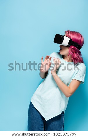 scared young woman with pink hair in vr headset on blue background