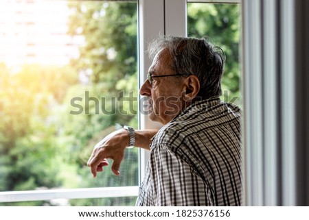 Shot of a senior man looking through window of his apartment. Portrait of a elder man in contemplation near window looking away and thinking. Pensive grey hair senior standing and relaxing, copy space