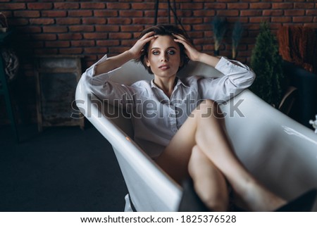 Portrait of a beautiful brunette lying in an empty bathroom holding legs up, wearing a white shirt and black shoes on red brick wall background.