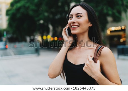Young freelancer female is having business phone conversation while walking down the street. A charming woman in trendy clothes is holding a mobile phone while standing on an urban background.