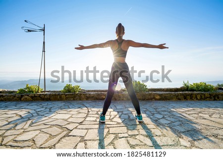 Fit muscular woman working out at sunrise on a stone patio facing the rising sun with arms outstretched outdoors in the garden in a health and fitness concept