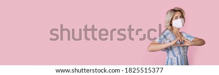 Cute blonde woman wearing a medical mask and dress is gesturing heart sign on a pink wall with free space