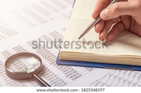 close up businessman writing and note on notebook with silver pen on table full of financial documents with digits and magnifying glass. banner