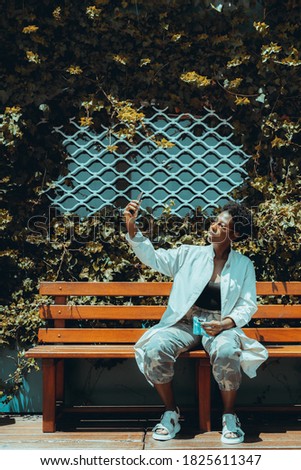 A young stylish black woman in sunglasses, white trench, and camouflage pants is sitting on a wooden bench near the window and the wall overgrown with ivy and making a selfie using her smartphone