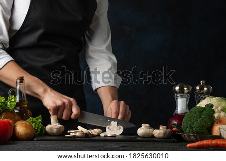 Close-up of the chef in black apron cuts with knife mushrooms on chopping black board at the professional restaurant kitchen on dark blue background. Backstage of preparing dish with vegetables.
