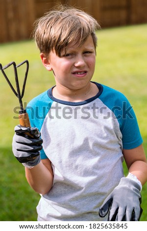 Vertical photo of a sad boy with a gardening fork in his hand not looking into the camera over the green grass background