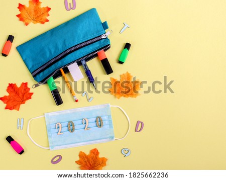 back to school concept.....blue pencil case with accessories, medical mask, alphabet, numbers, 2020 and autumn leaves lies on the left on a yellow background with a place for text on the right, top vi