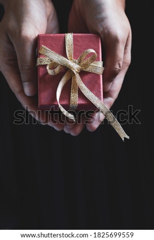 Close up of selective focus on red gift box tied with gold ribbon holds by hands with blurry noise black background